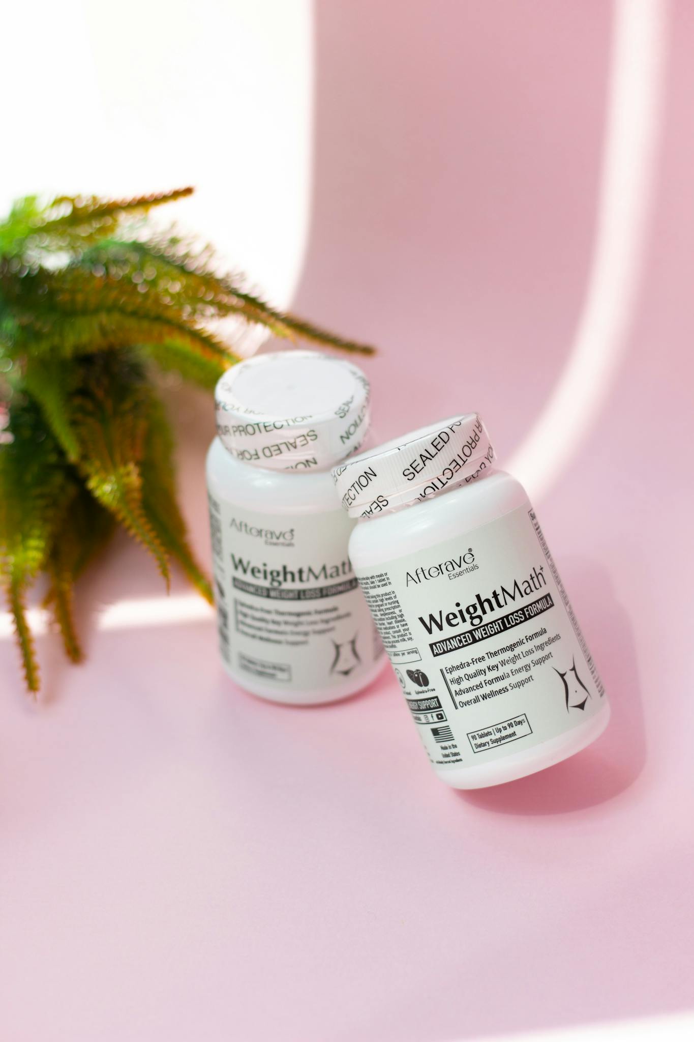 Two weight management supplement bottles on a pink surface with greenery.